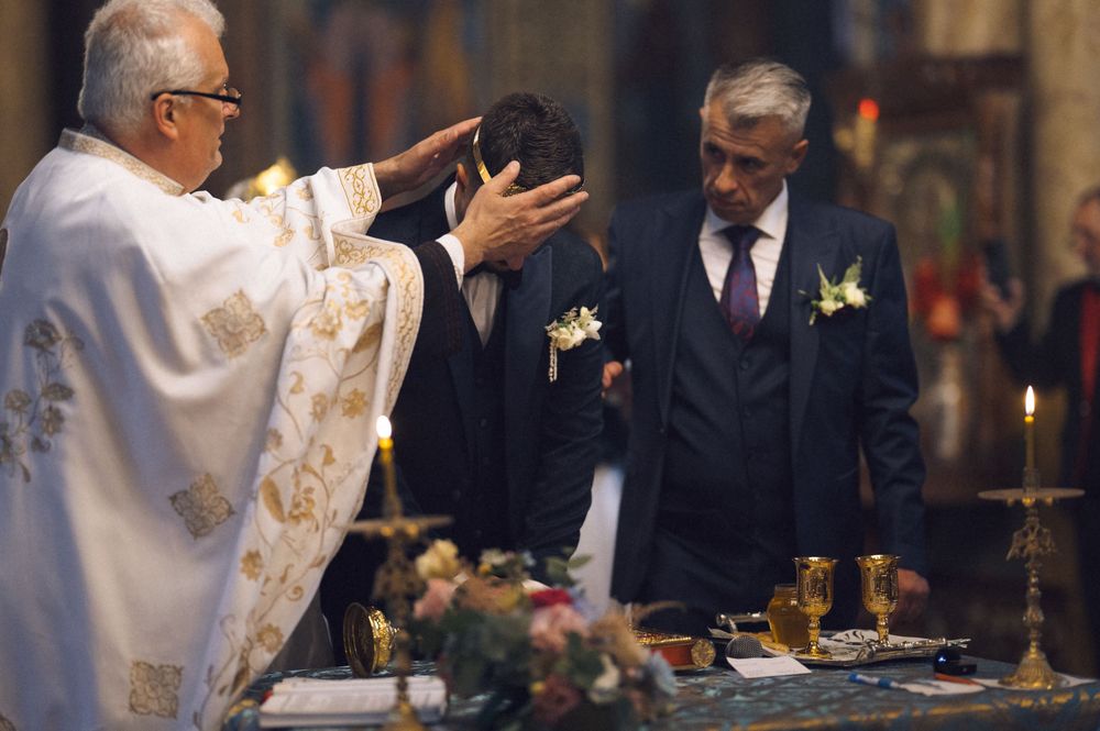 Orthodox priest blessing groom during religious wedding ceremony in ornate church with candles and floral decorations.