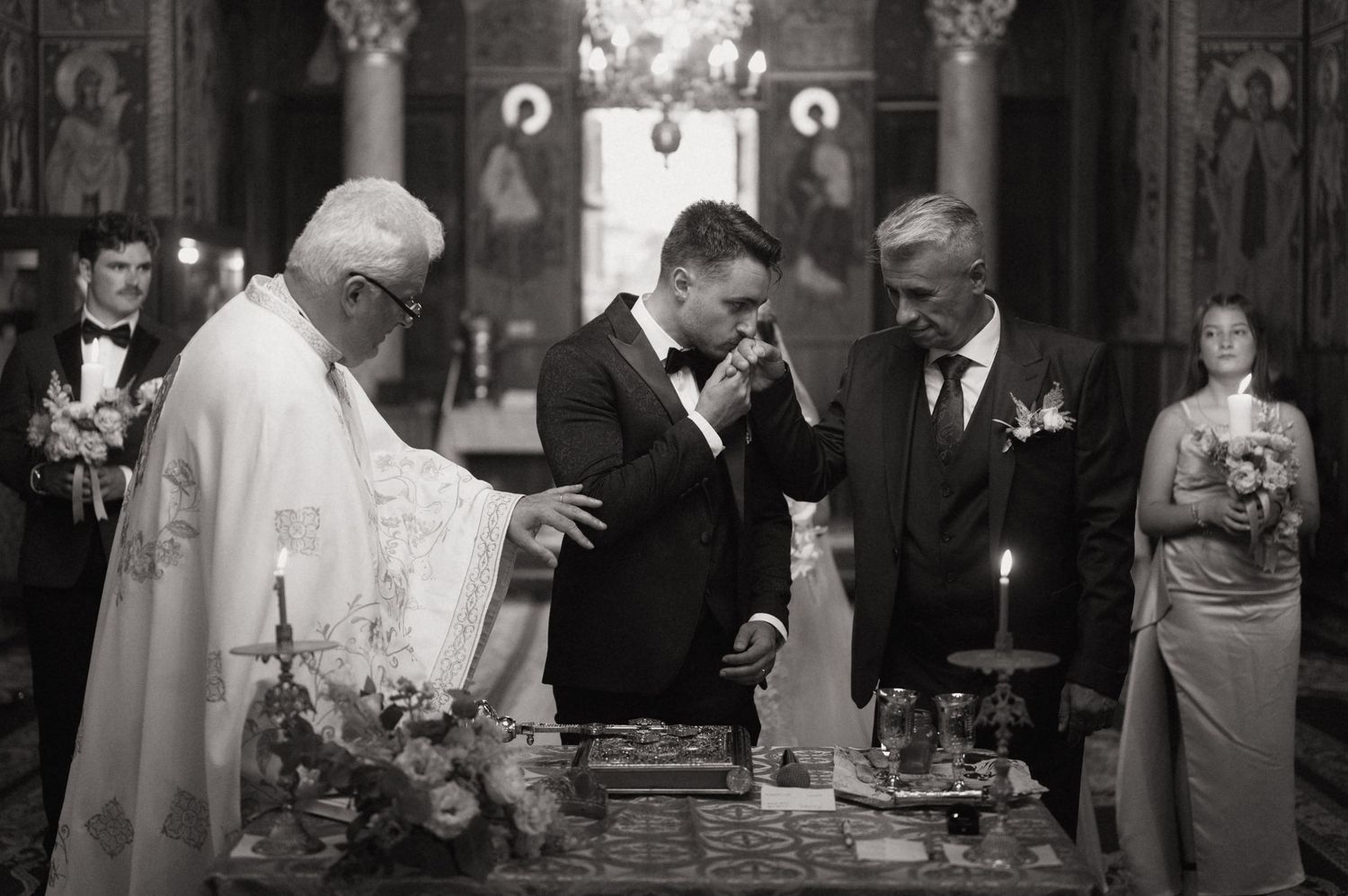 Black and white photo of a groom kissing the hand of a priest during an Orthodox wedding ceremony.