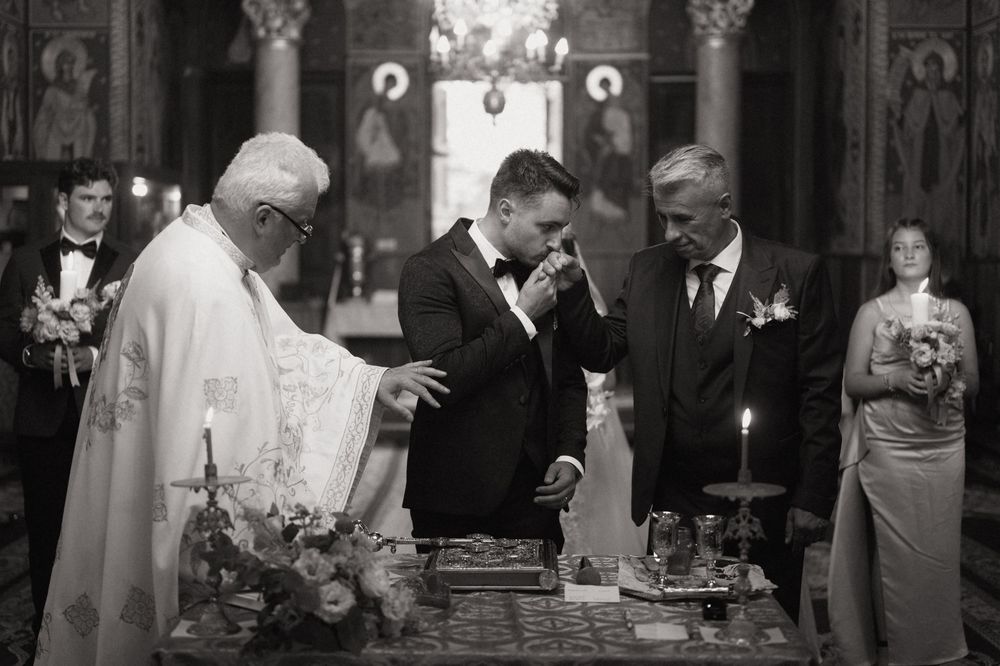 Black and white photo of a groom kissing the hand of a priest during an Orthodox wedding ceremony.