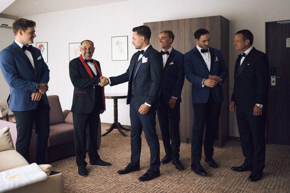 Six men in formal suits and tuxedos gather in a hotel room during wedding preparations, with the groom receiving congratulations.