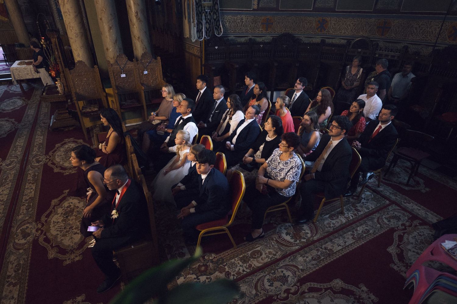 Aerial view of elegantly dressed guests seated in ornate chairs at a formal indoor ceremony or event.