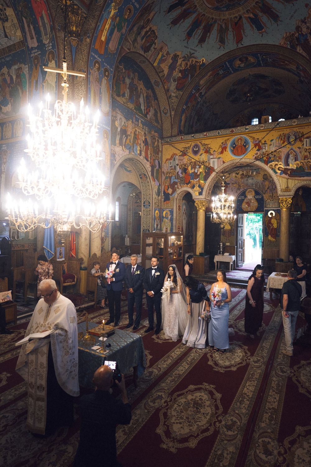 Orthodox church wedding ceremony with bride and groom standing before priest, ornate frescoes and chandelier in background.