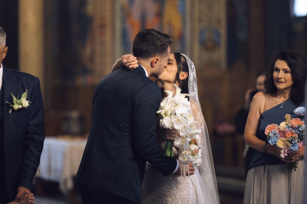 Bride and groom share first kiss at church wedding altar, bridesmaid holding floral bouquet in background.