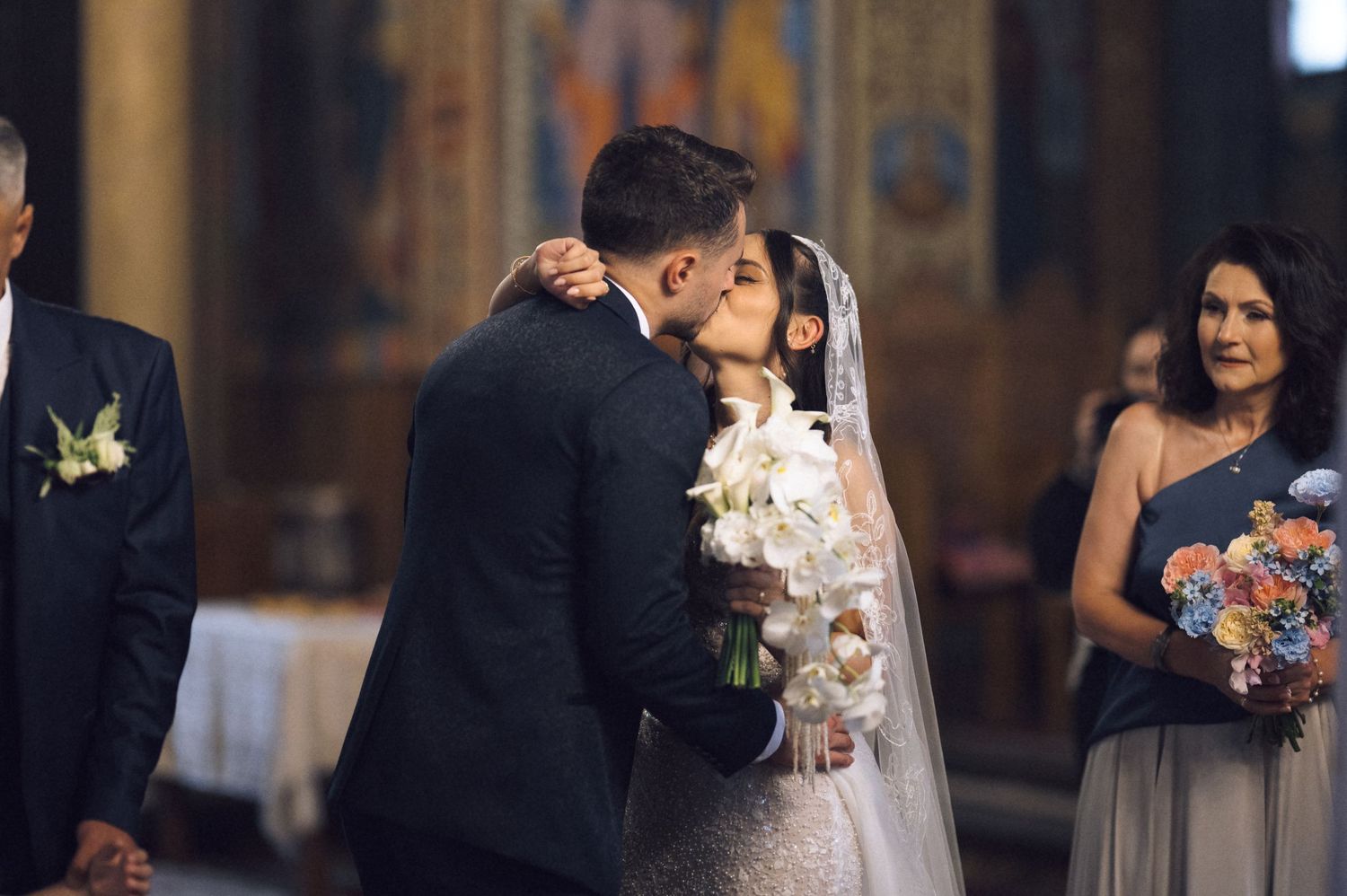 Bride and groom share first kiss at church wedding altar, bridesmaid holding floral bouquet in background.