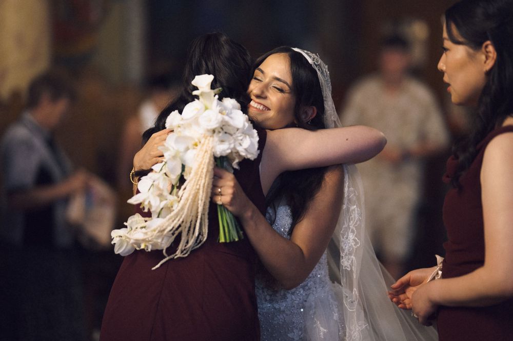 Bride in white gown hugging a guest while holding a bouquet of white flowers at a wedding ceremony.