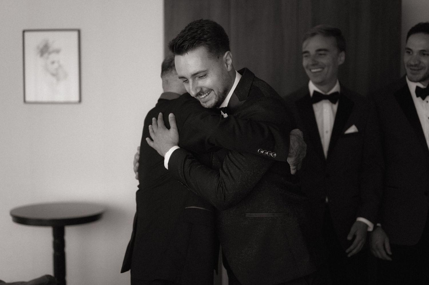 Men in tuxedos sharing a warm hug at a formal event, captured in black and white photography.