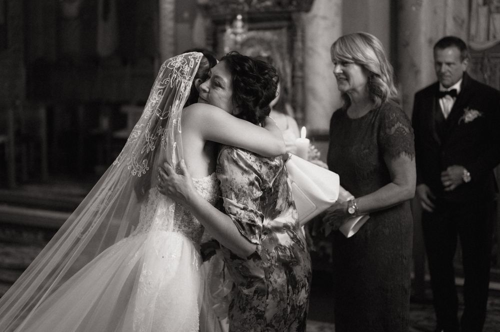 Black and white wedding photo of bride embracing guest in church, with woman in floral dress and man in suit nearby.