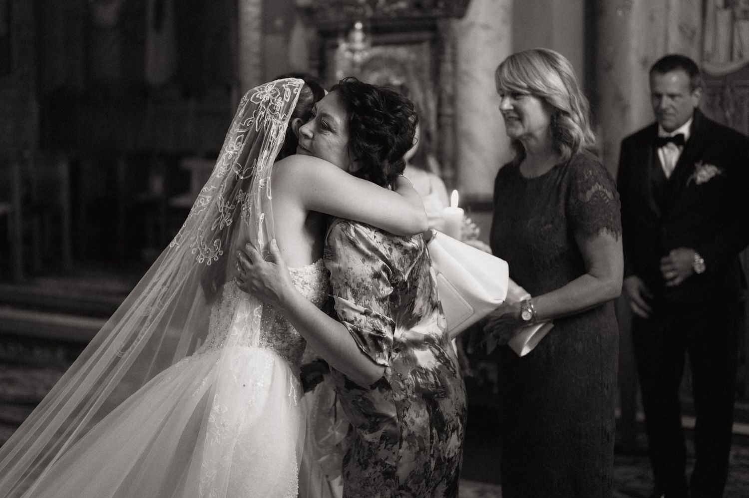 Black and white wedding photo of bride embracing guest in church, with woman in floral dress and man in suit nearby.