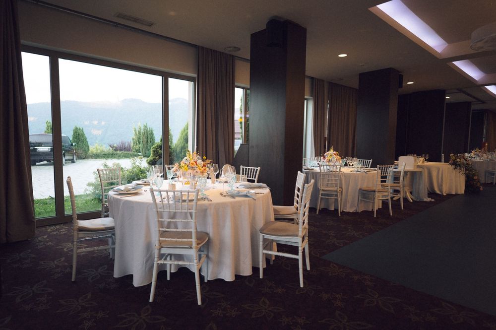 Elegant banquet hall with white-draped round tables, chiavari chairs, and panoramic mountain views through floor-to-ceiling windows.