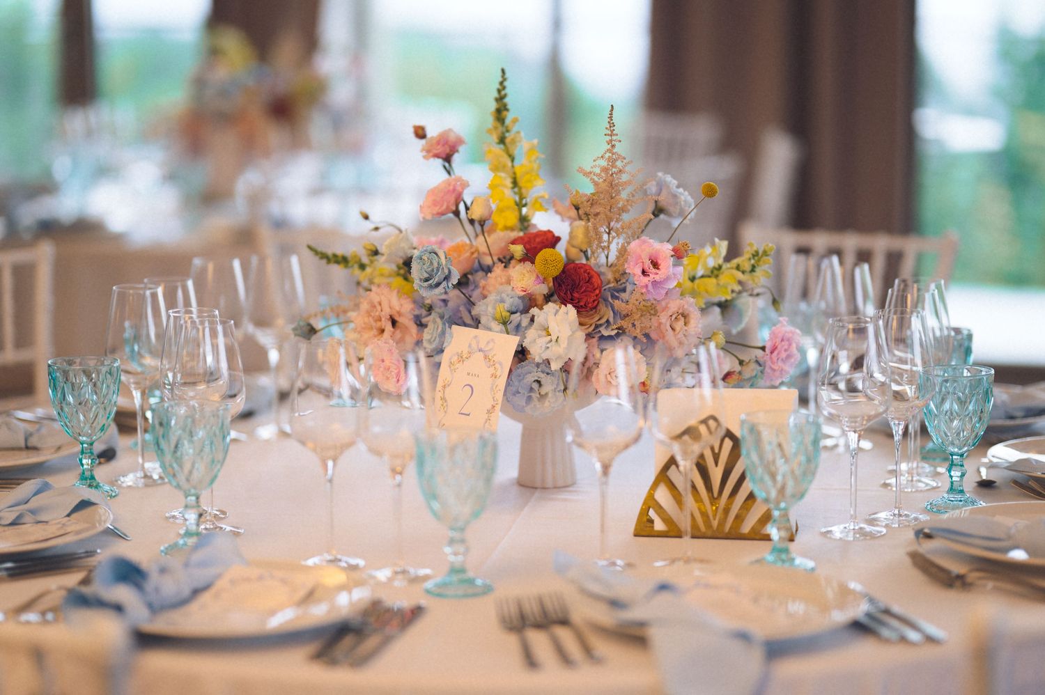 Wedding reception table set with blue glassware, gold accents, and colorful floral centerpiece.