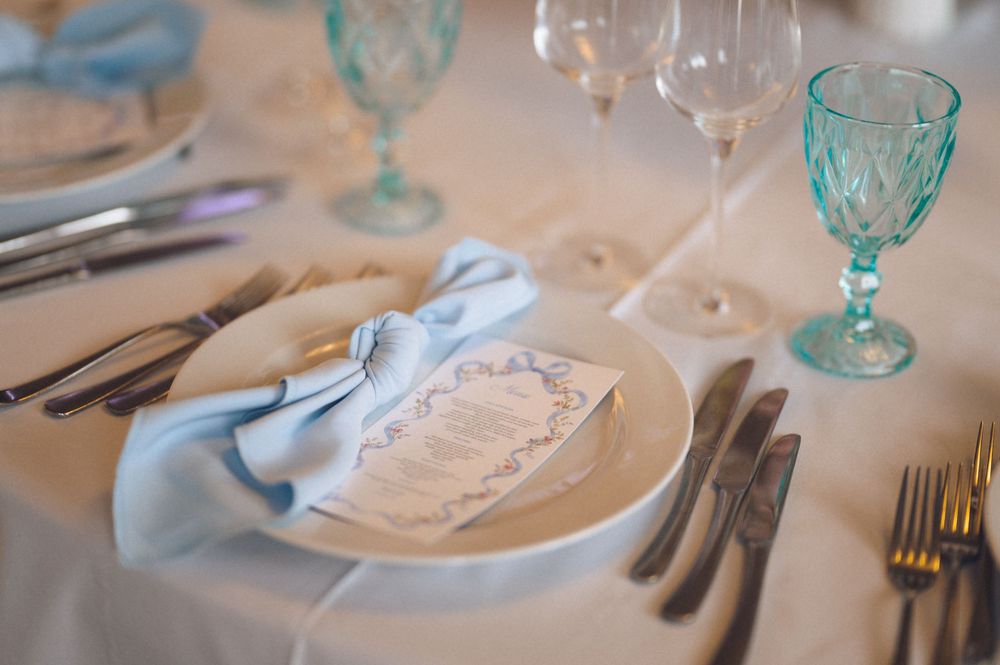 Elegant wedding table setting with blue napkin, white plate, menu card, crystal glasses, and silverware on white tablecloth.