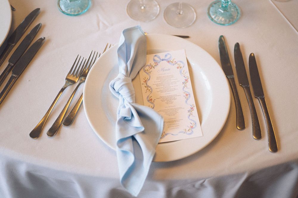 Elegant wedding table setting with blue napkin, white plate, menu card, and gold silverware on a white tablecloth.