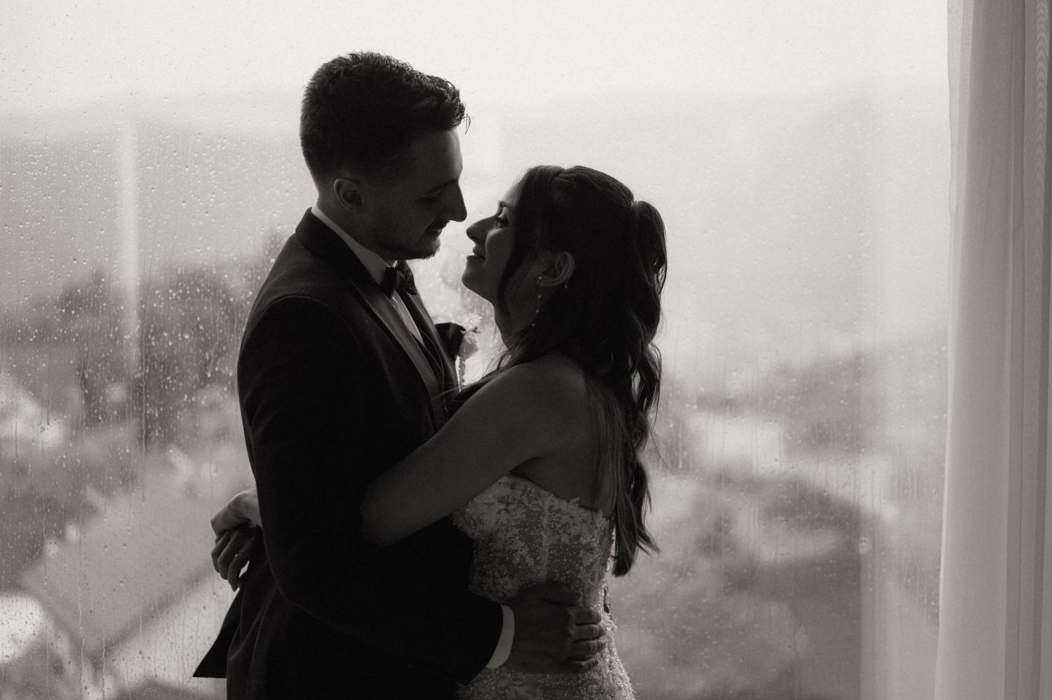 Black and white photo of a couple sharing a romantic embrace near a rain-streaked window on their wedding day.