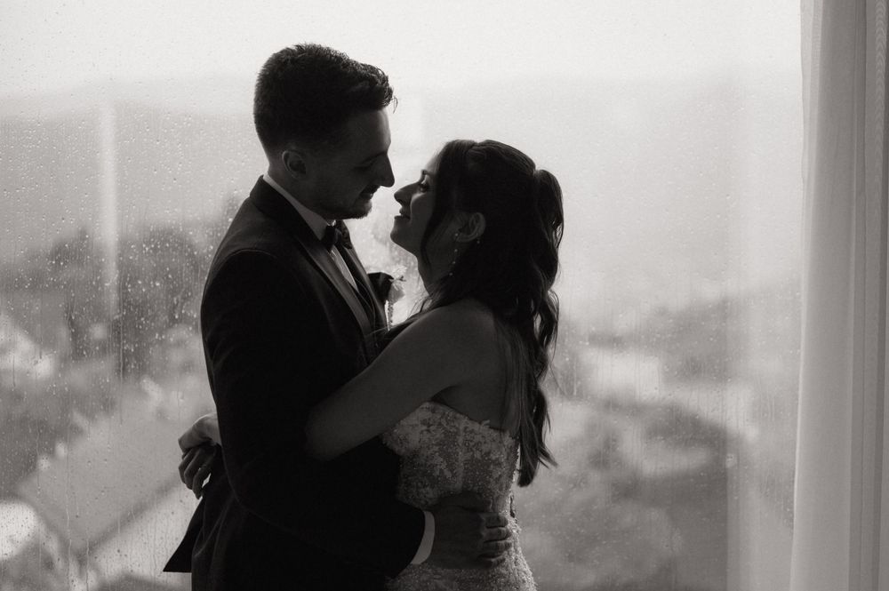 Black and white photo of a couple sharing a romantic embrace near a rain-streaked window on their wedding day.