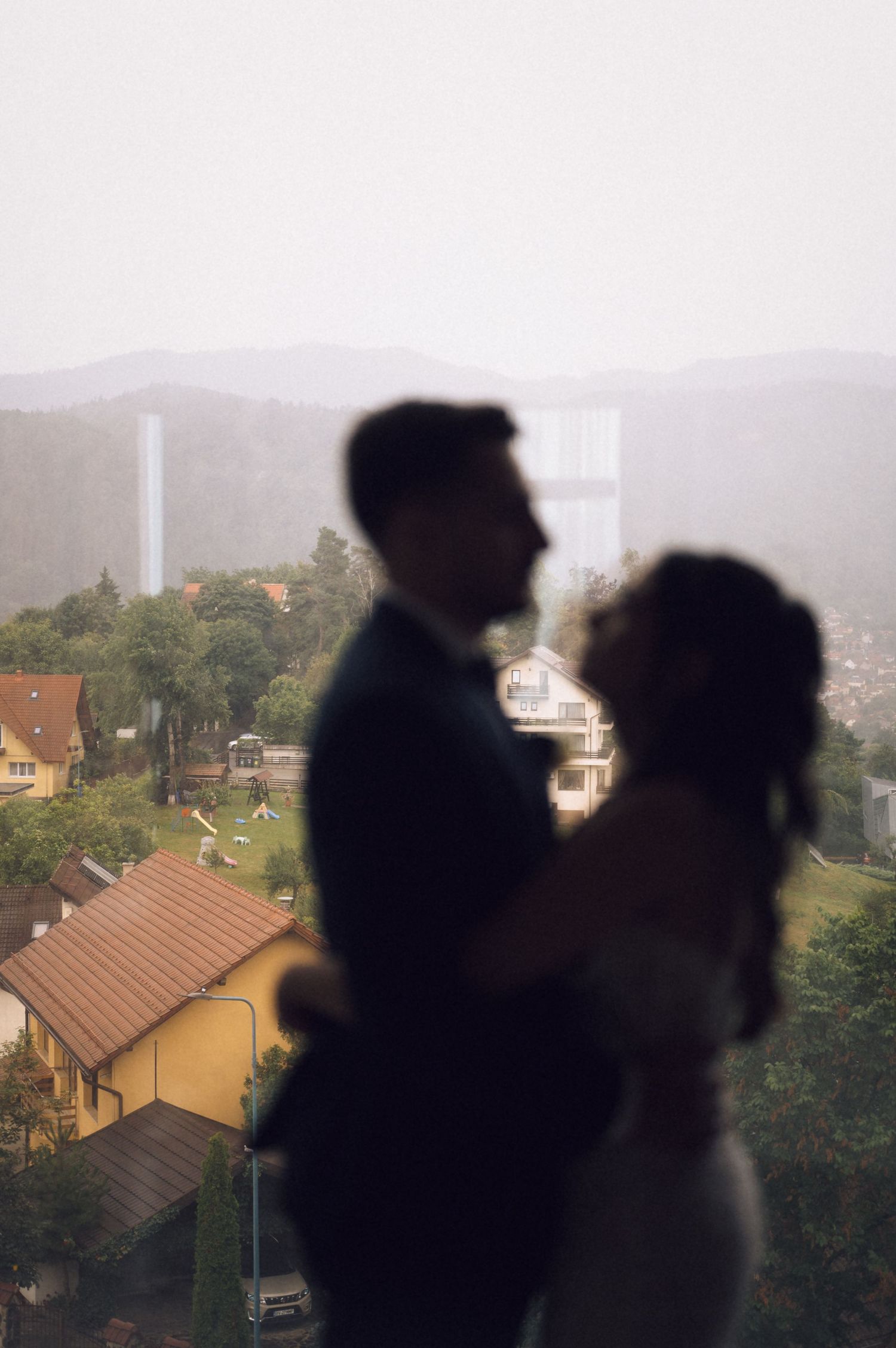 Silhouette of a couple embracing near a window with a misty town and mountains in the background.