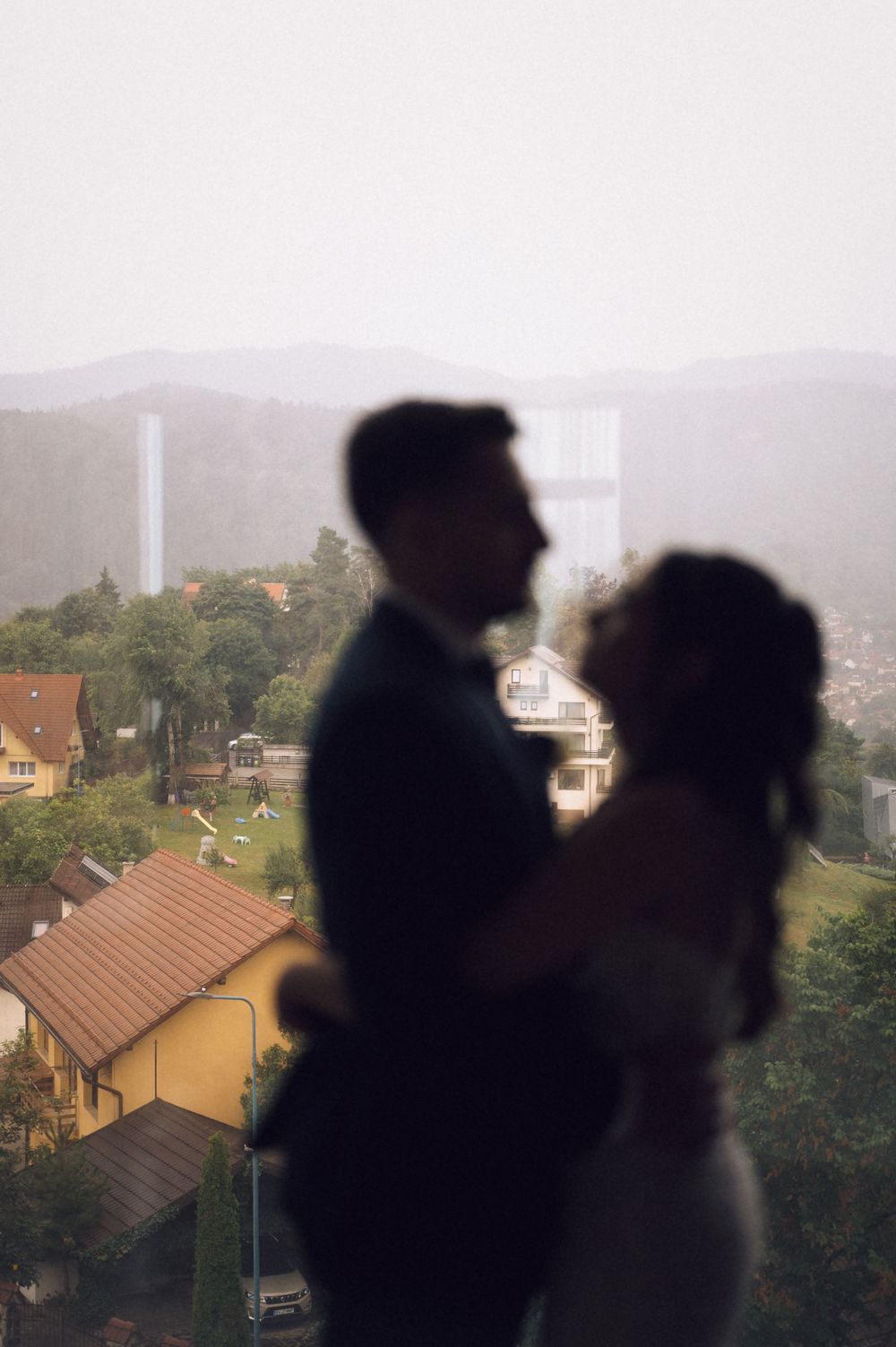 Silhouette of a couple embracing near a window with a misty town and mountains in the background.