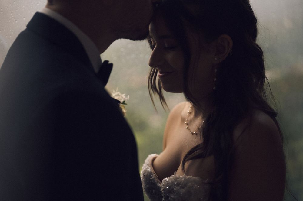 Intimate wedding couple portrait in moody lighting, bride in lace gown with pearl earrings, groom in dark suit with boutonniere.