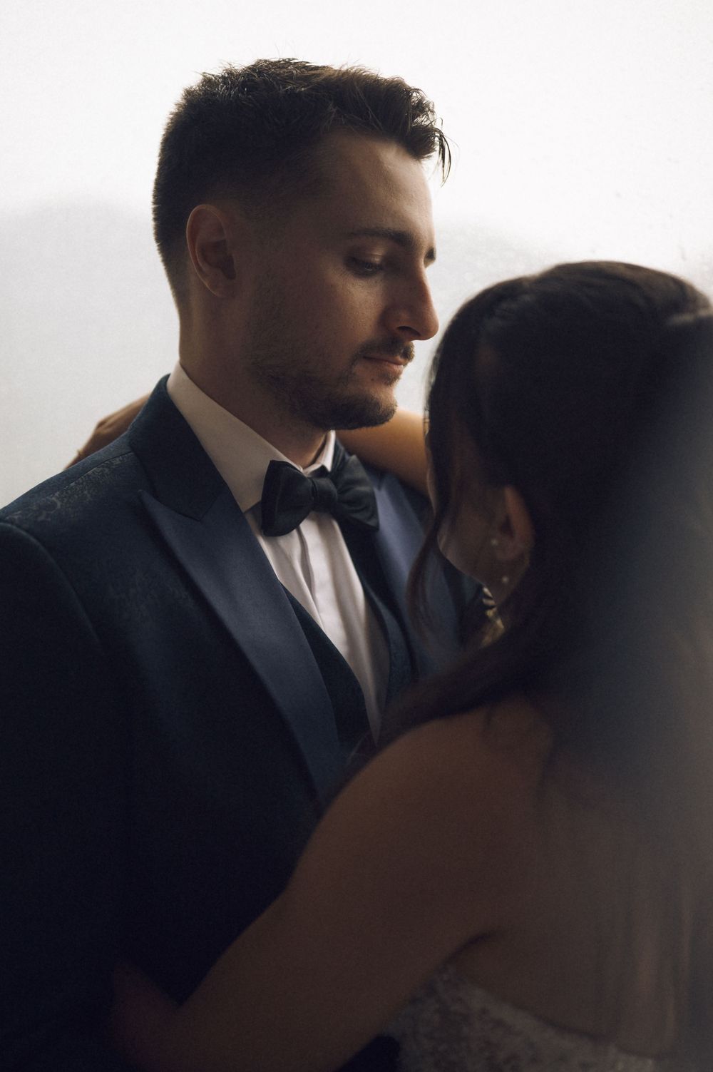 A groom in a navy tuxedo with bow tie gazes down at his bride during an intimate wedding moment.