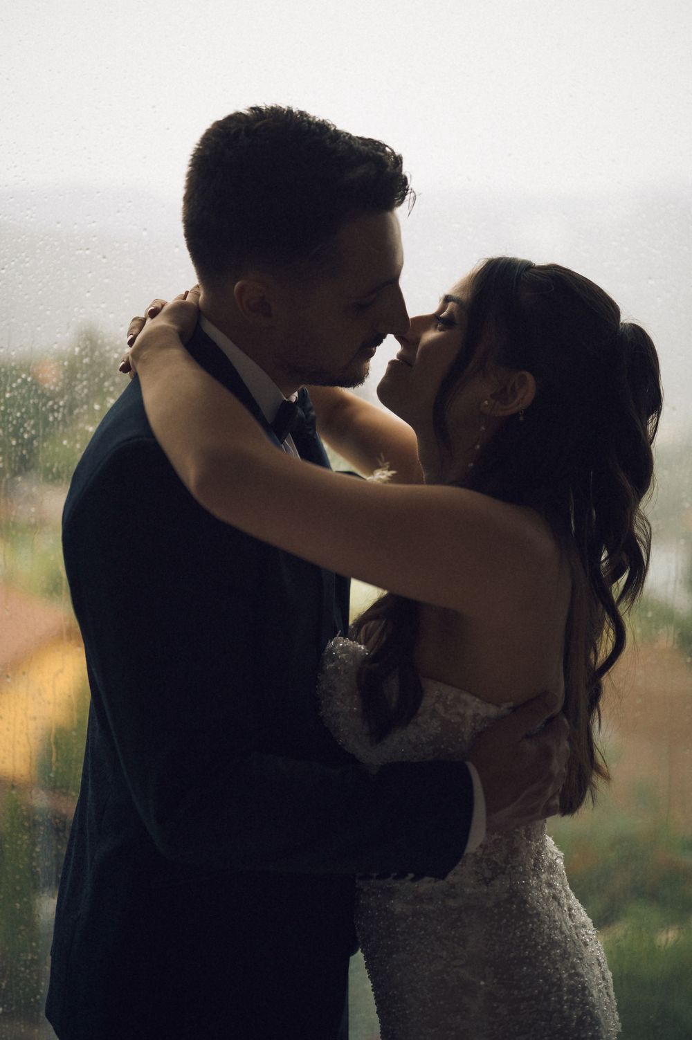 A bride and groom share a romantic almost-kiss embrace in wedding attire against a rain-streaked window backdrop.