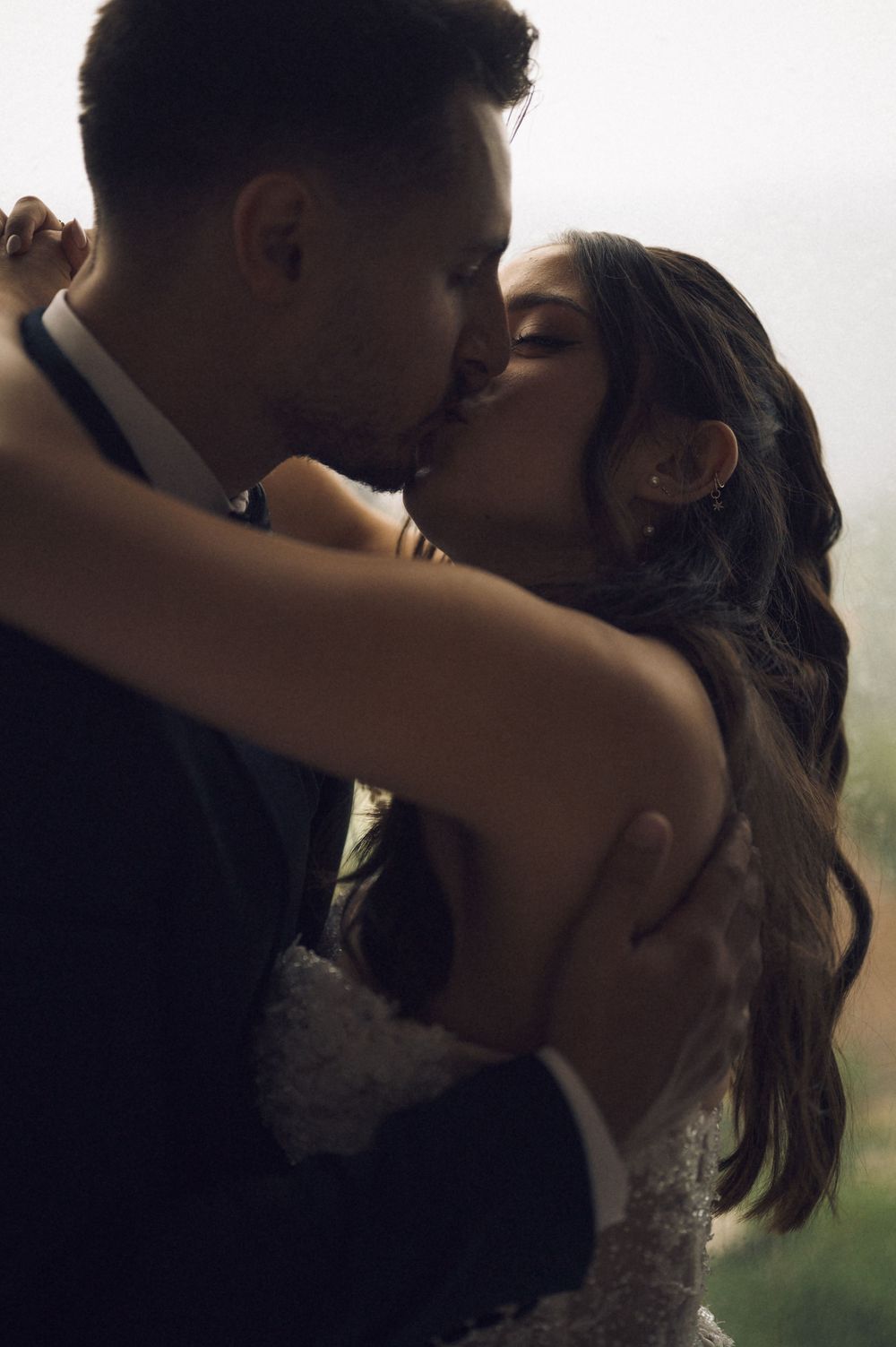 A bride and groom share a romantic kiss in soft light, dressed in wedding attire.