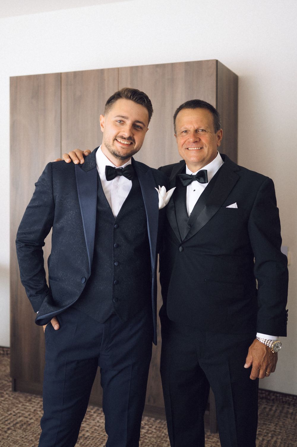 Two men in black tuxedos with bow ties pose together smiling at a formal event.