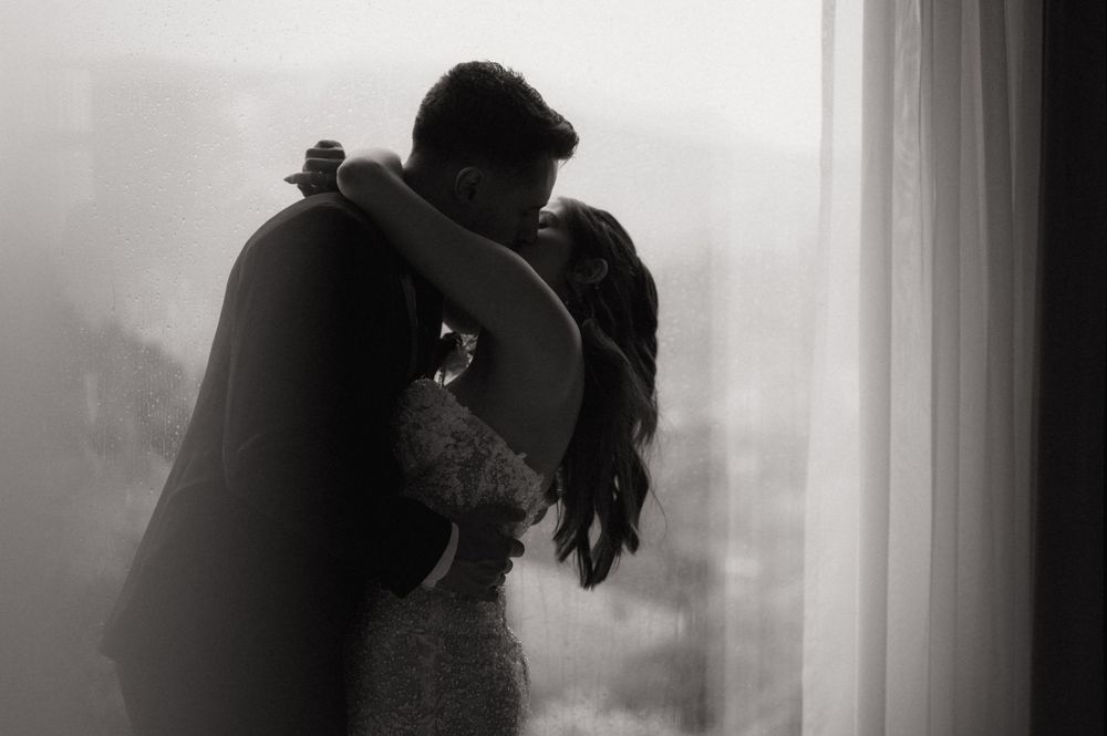 Black and white photo of a couple embracing near a window, sharing a romantic moment in wedding attire.