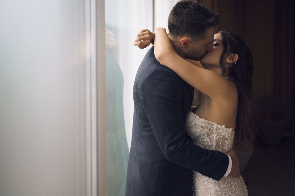 Bride and groom sharing a romantic kiss near a window on their wedding day.