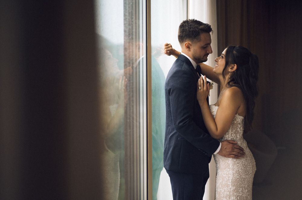 A couple in wedding attire shares a romantic moment by a sunlit window, the bride in lace and groom in a dark suit.