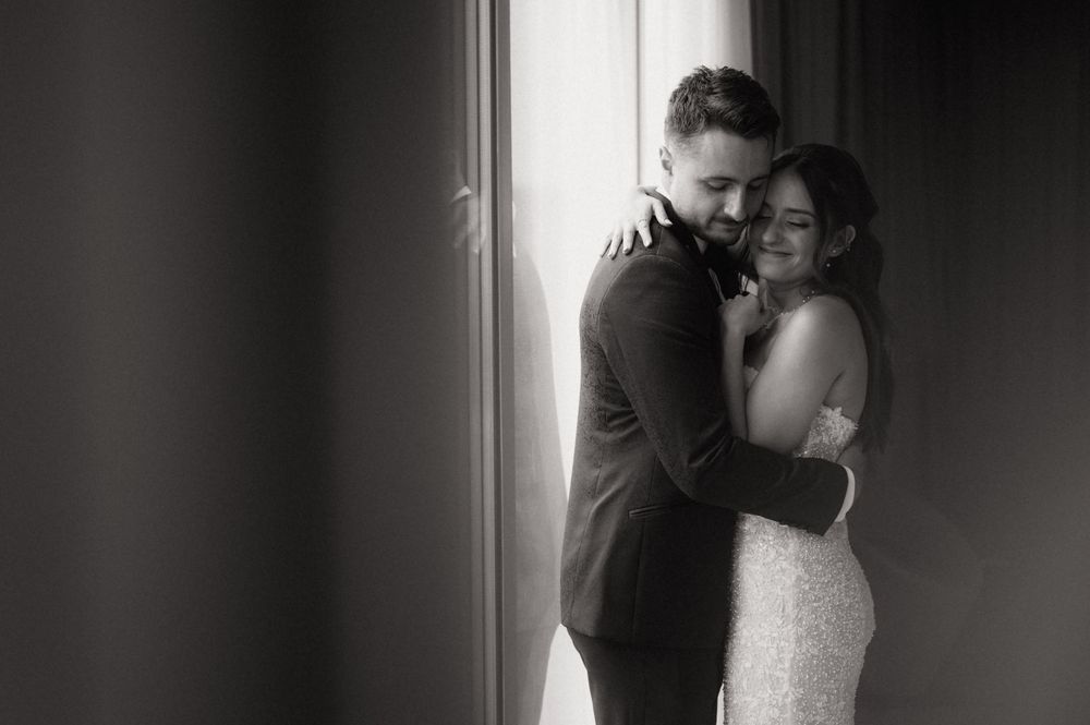 Black and white wedding photo of bride and groom embracing tenderly by a window in elegant attire.