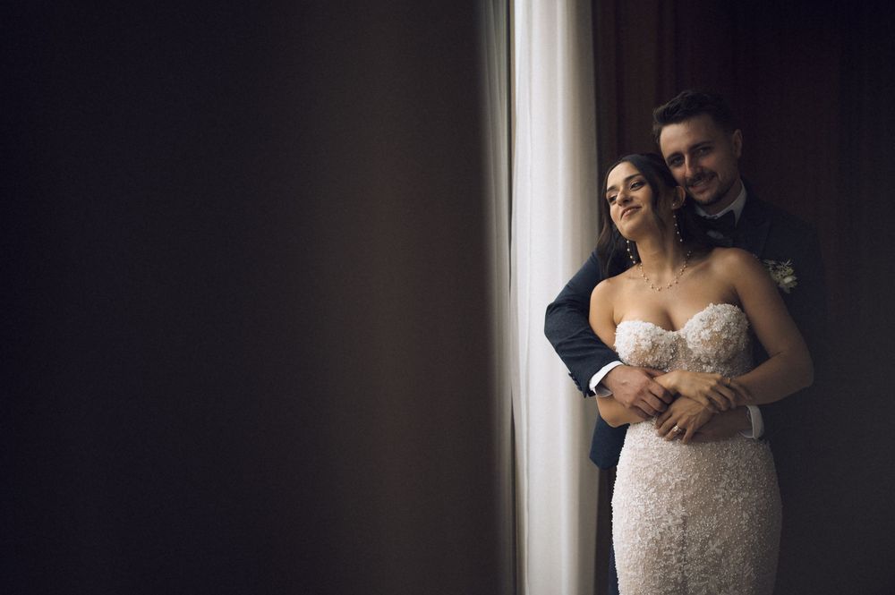 A bride and groom share a tender embrace near a window, the bride in a lace gown and groom in a dark suit.