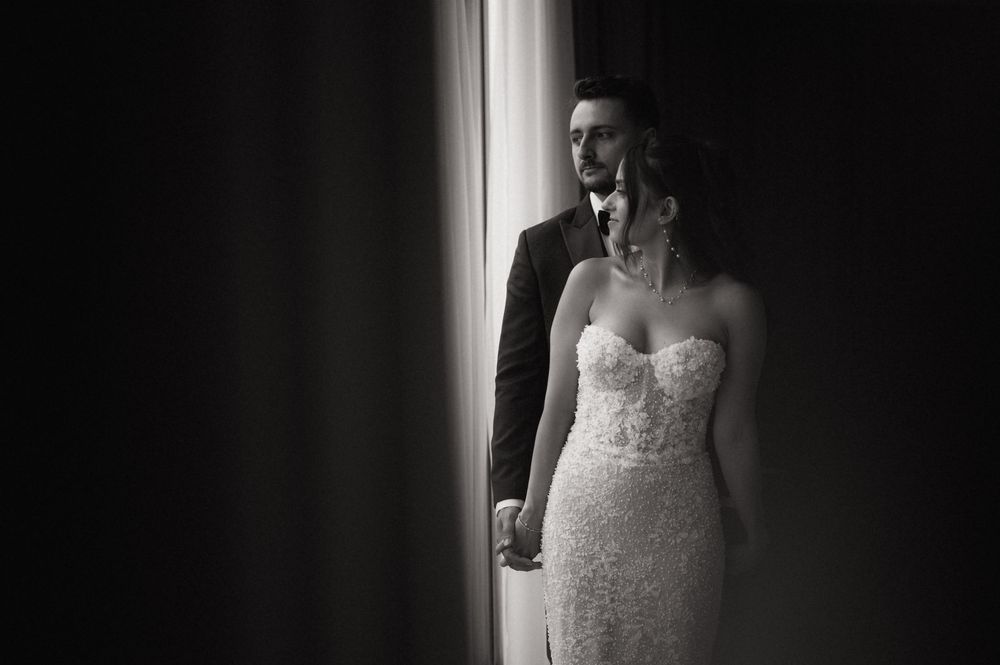 Black and white wedding photo of bride in lace gown and groom in suit standing by a bright window.