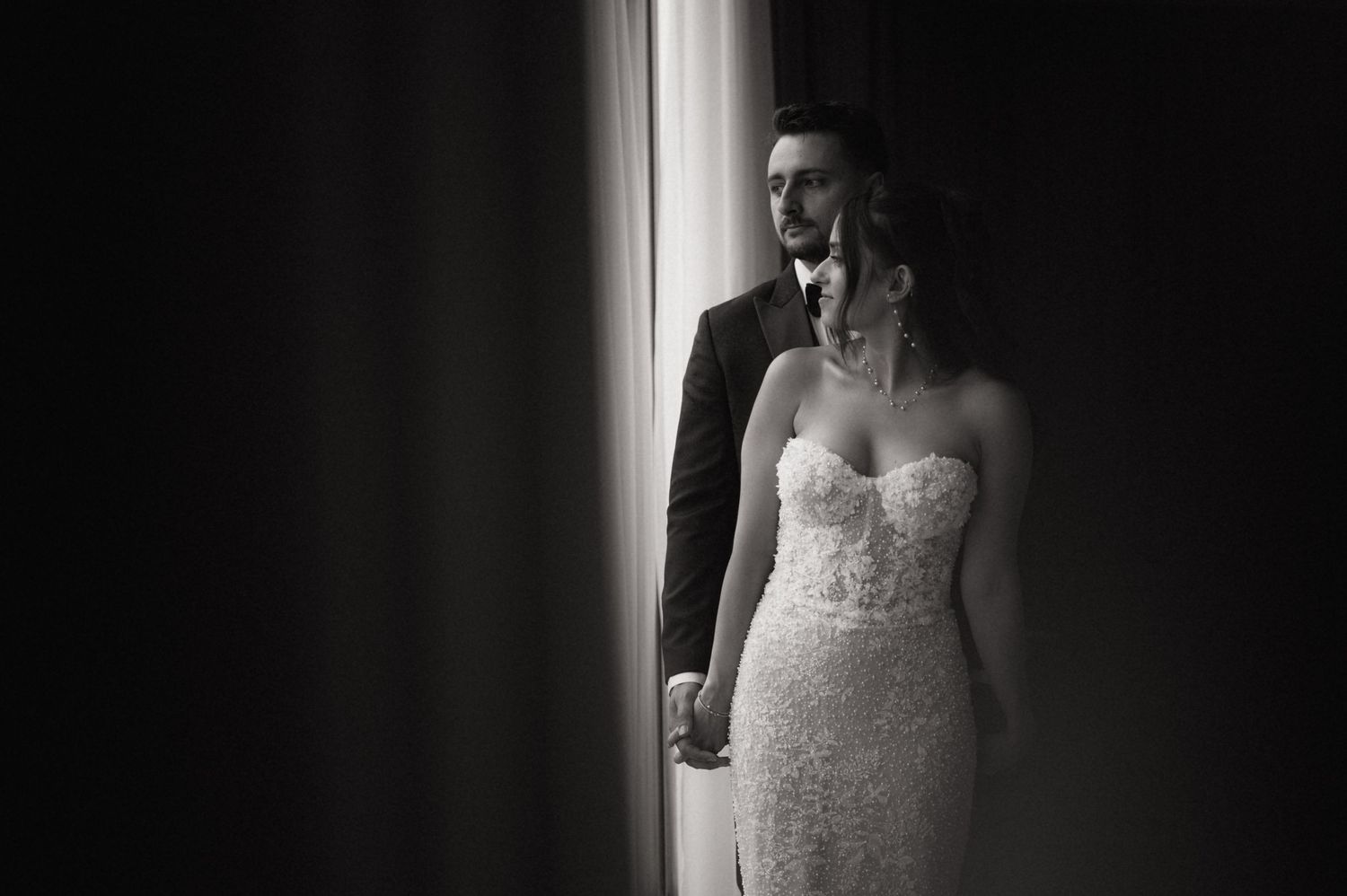 Black and white wedding photo of bride in lace gown and groom in suit standing by a bright window.