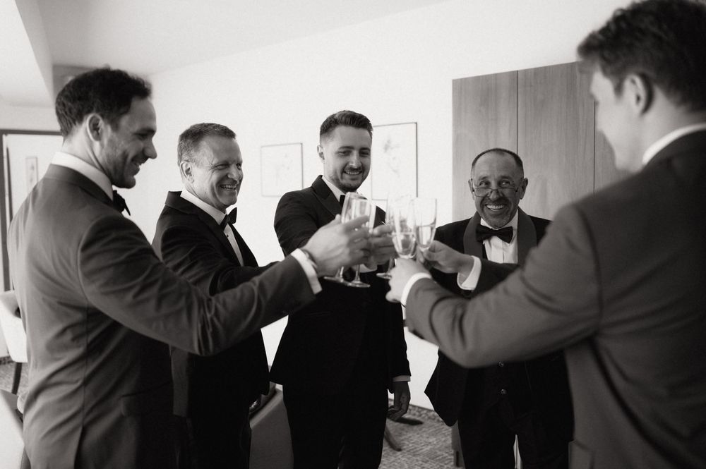 Groomsmen in suits toasting with champagne glasses before a wedding ceremony in black and white photo.