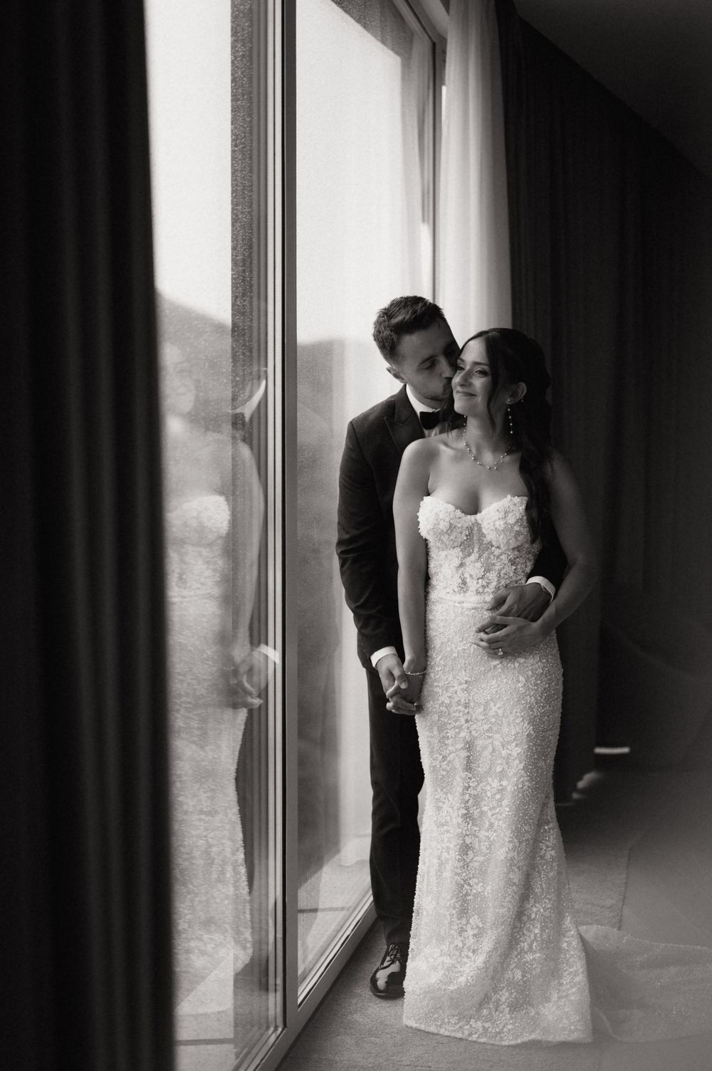 Black and white photo of a bride and groom holding hands by large windows, groom kissing bride's head in elegant wedding attire.