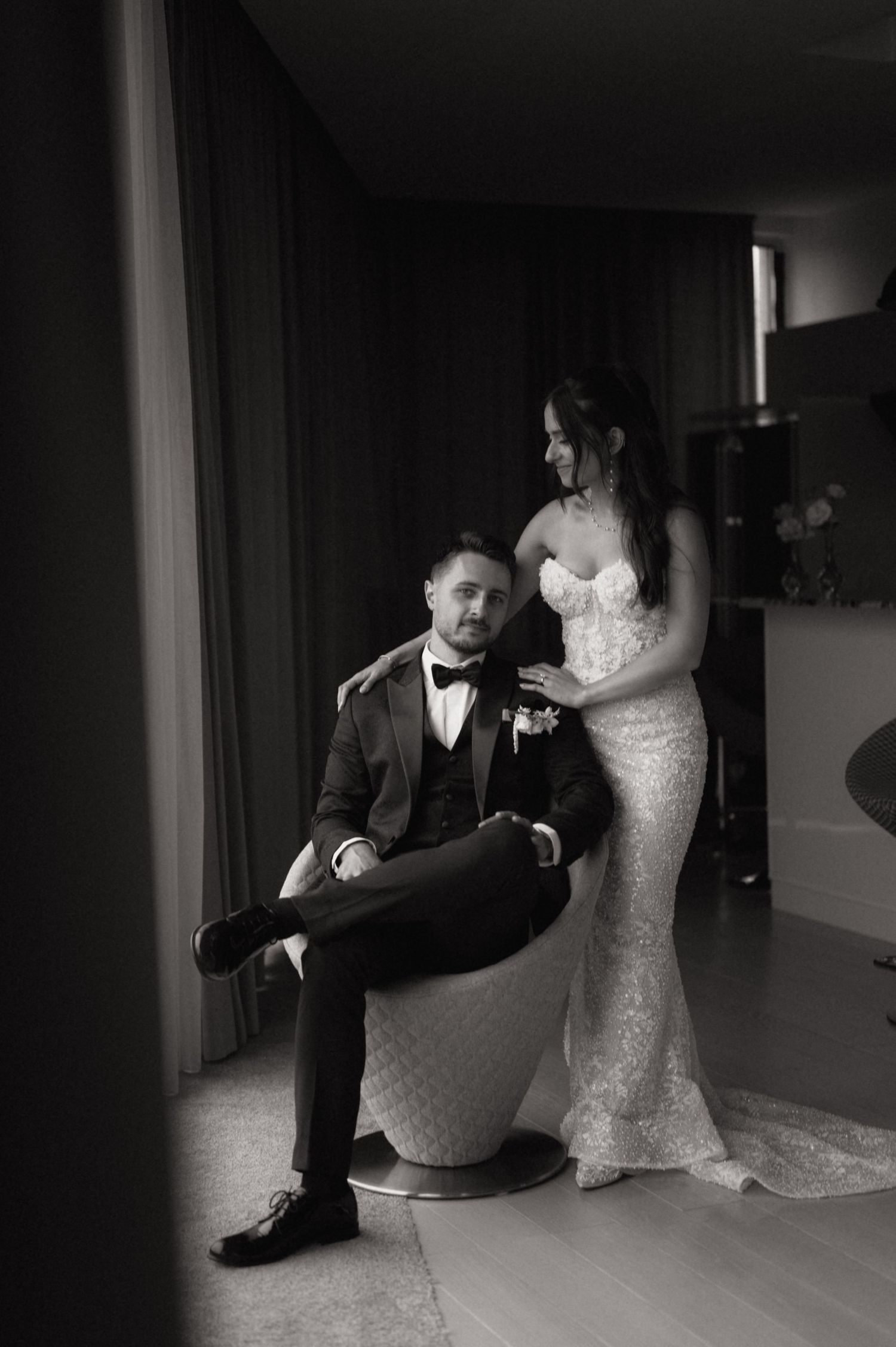 Black and white photo of a bride in a lace gown standing beside a seated groom in a tuxedo at their wedding.