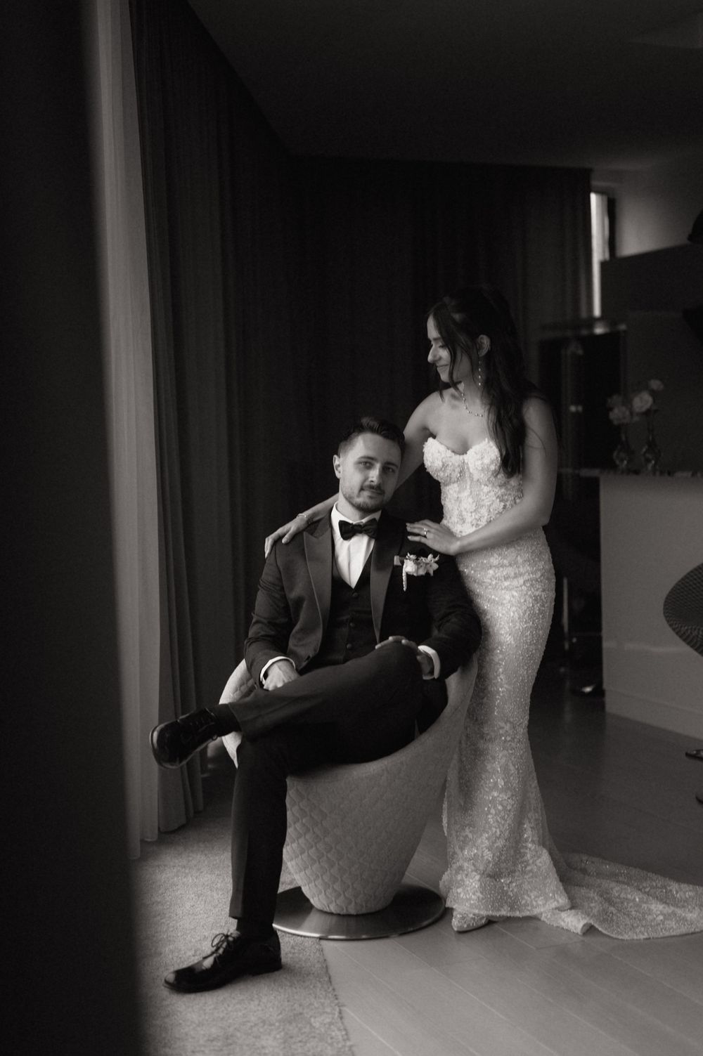 Black and white photo of a bride in a lace gown standing beside a seated groom in a tuxedo at their wedding.