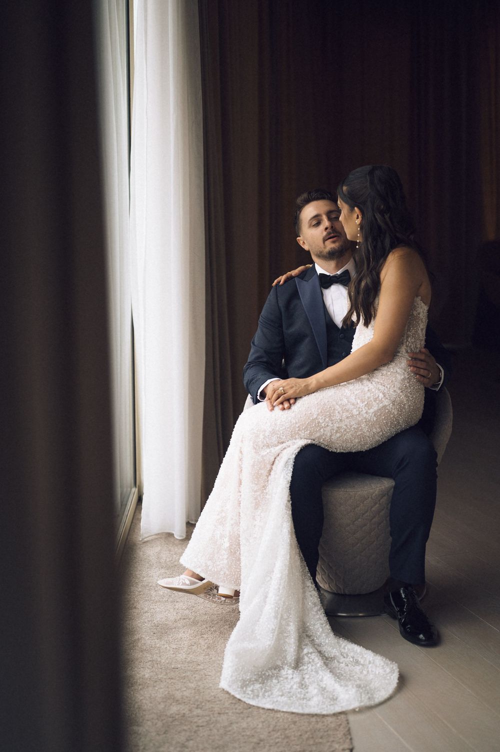 Bride in lace gown embraces groom in tuxedo seated by window in romantic wedding portrait.