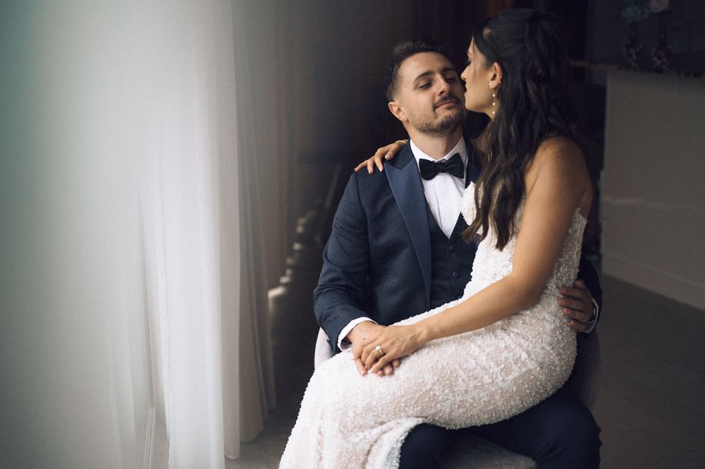 A bride in a white lace gown embraces a groom in a navy tuxedo as they sit together by a window.