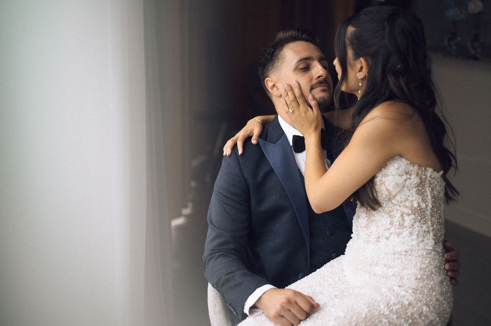 Bride in lace wedding dress tenderly holds groom's face as they share an intimate moment by a window.