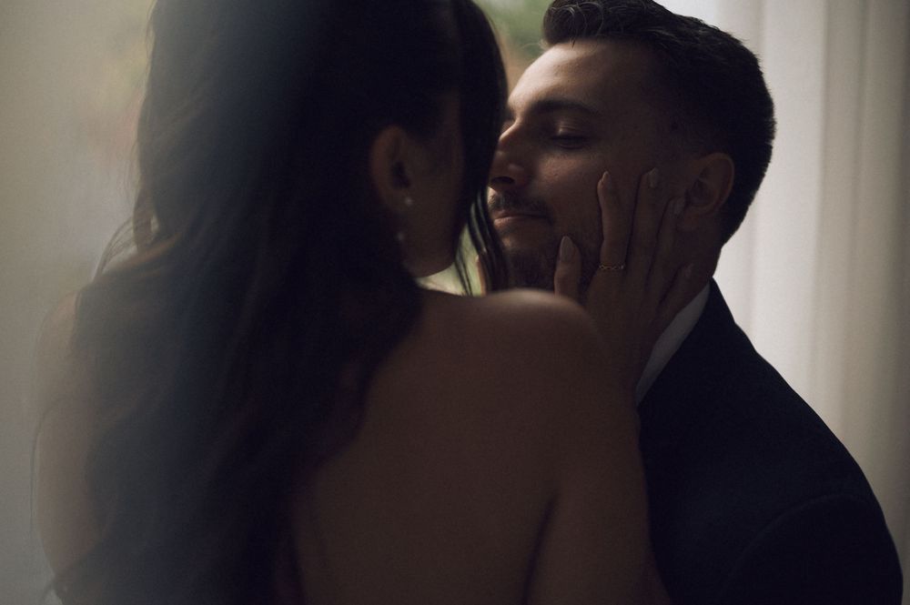 Couple shares a romantic near-kiss moment, bride's hand gently cupping groom's face in soft light.