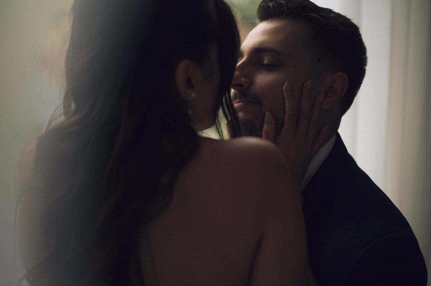 Couple shares a romantic near-kiss moment, bride's hand gently cupping groom's face in soft light.