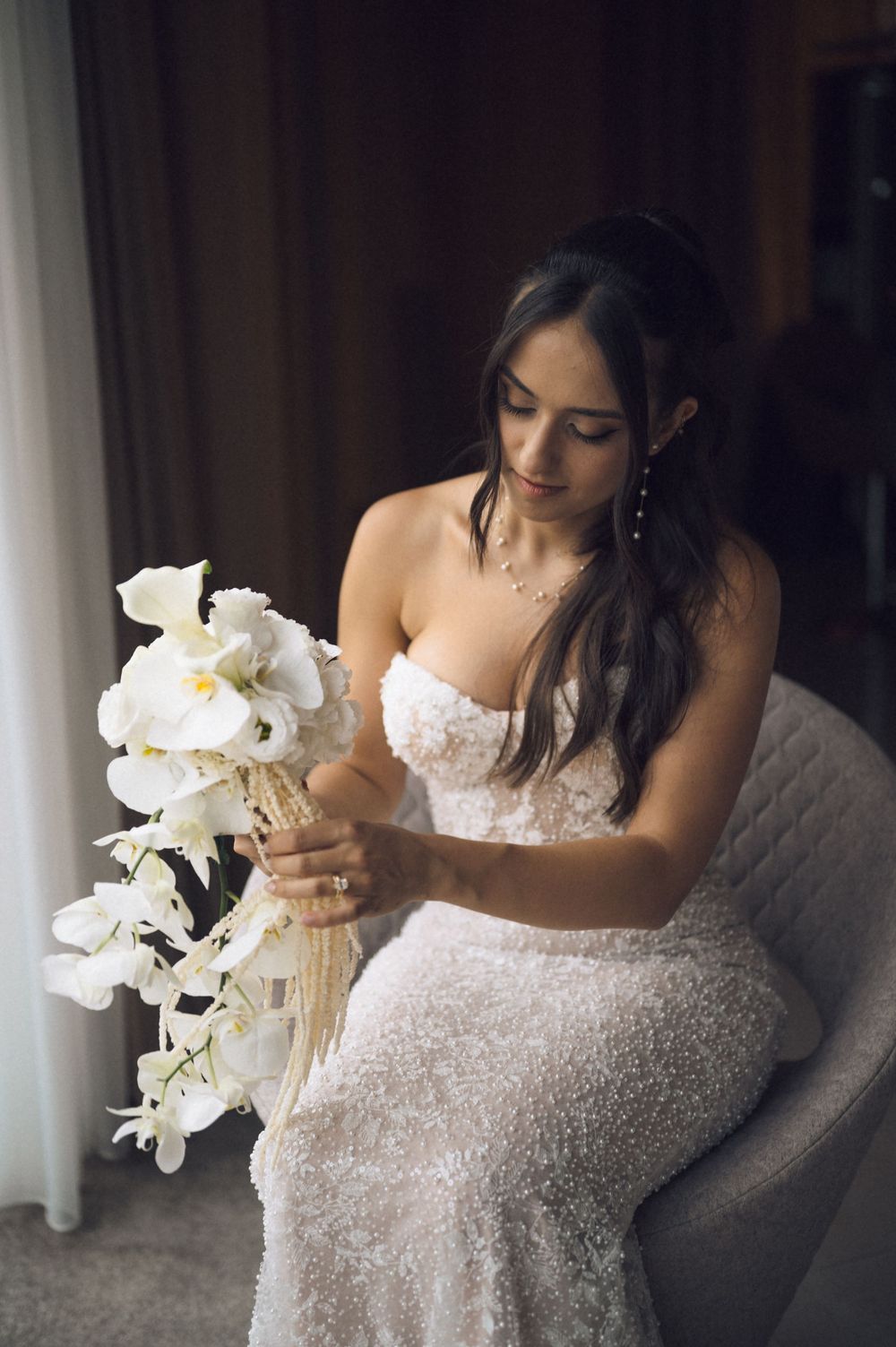 Bride in strapless lace gown sits holding white orchid bouquet by window soft natural light elegant wedding portrait.