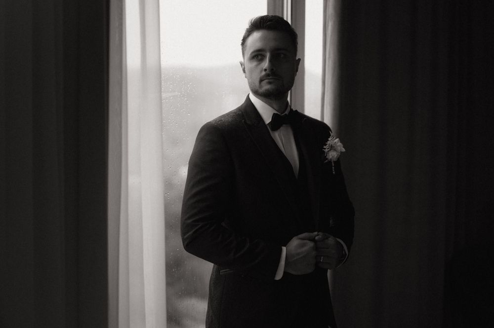 Black and white photo of a groom in a tuxedo with boutonniere adjusting his jacket by a window.