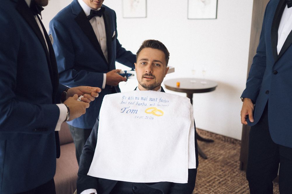 Groom seated in chair wearing a wedding bib, surrounded by groomsmen in navy suits during pre-wedding preparations.