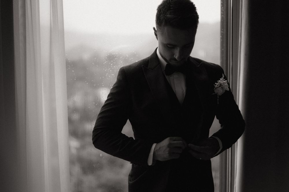 Black and white photo of a groom in a tuxedo adjusting his cufflinks by a window on his wedding day.