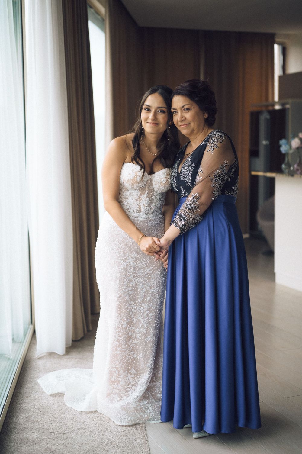 Bride in white lace gown poses with woman in blue evening dress near window in elegant indoor setting.