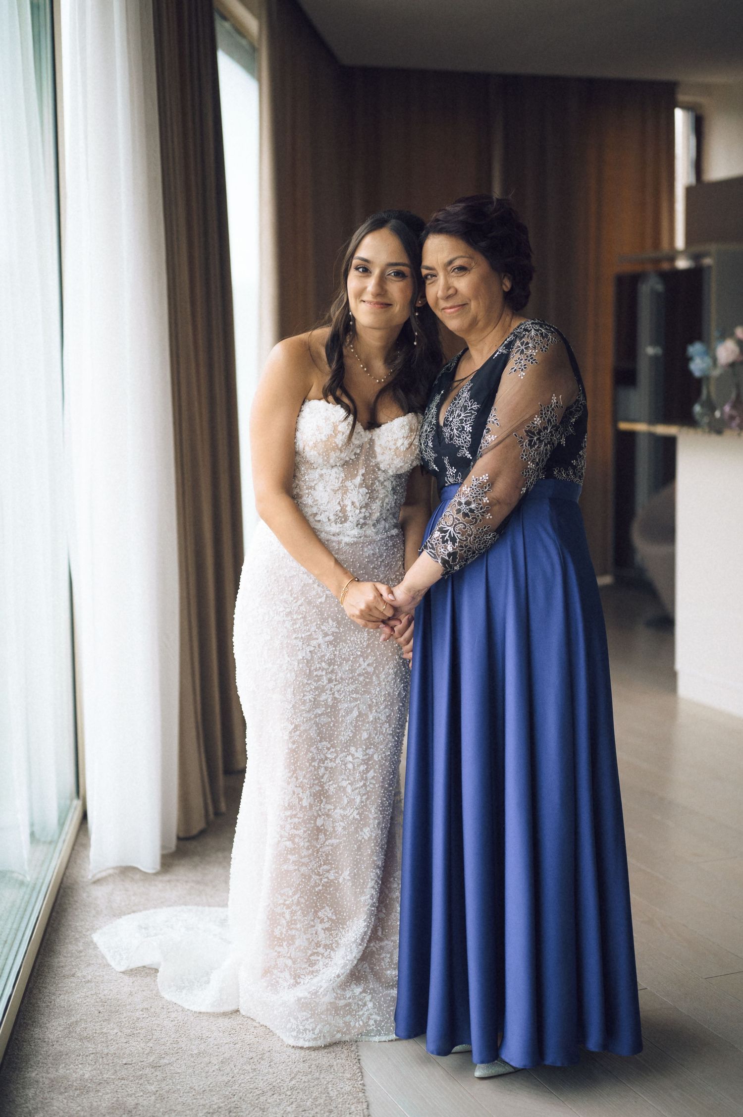 Bride in white lace gown poses with woman in blue evening dress near window in elegant indoor setting.