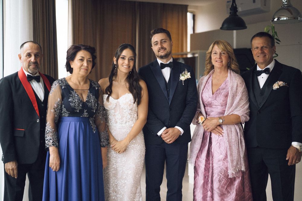 A wedding group photo featuring bride in white gown, groom in tuxedo, and four family members in formal attire.
