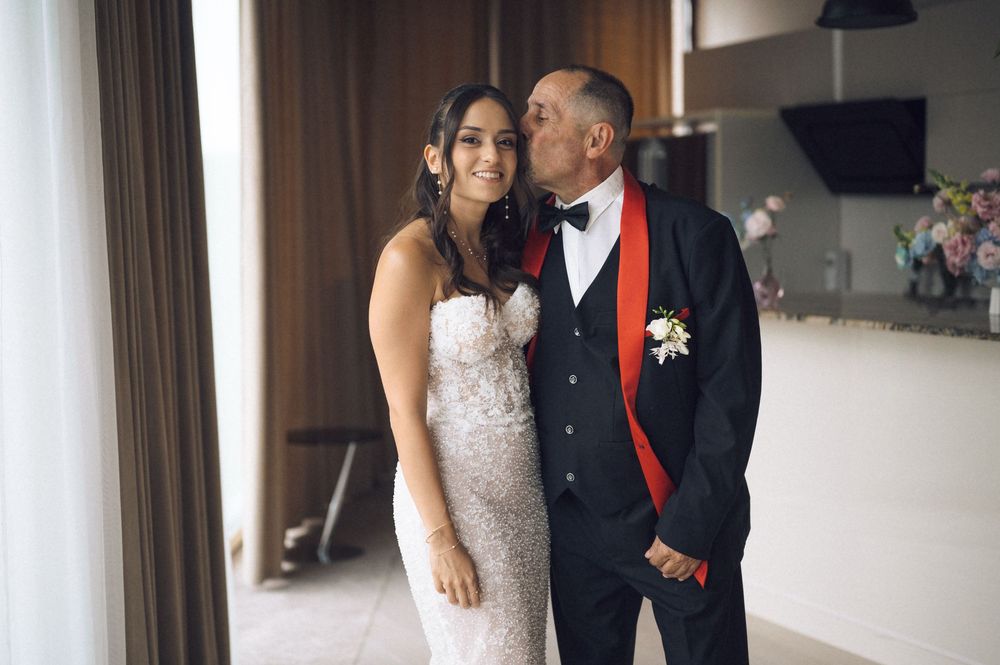 Groom in black tuxedo with red tie kisses bride in white lace wedding dress indoors.