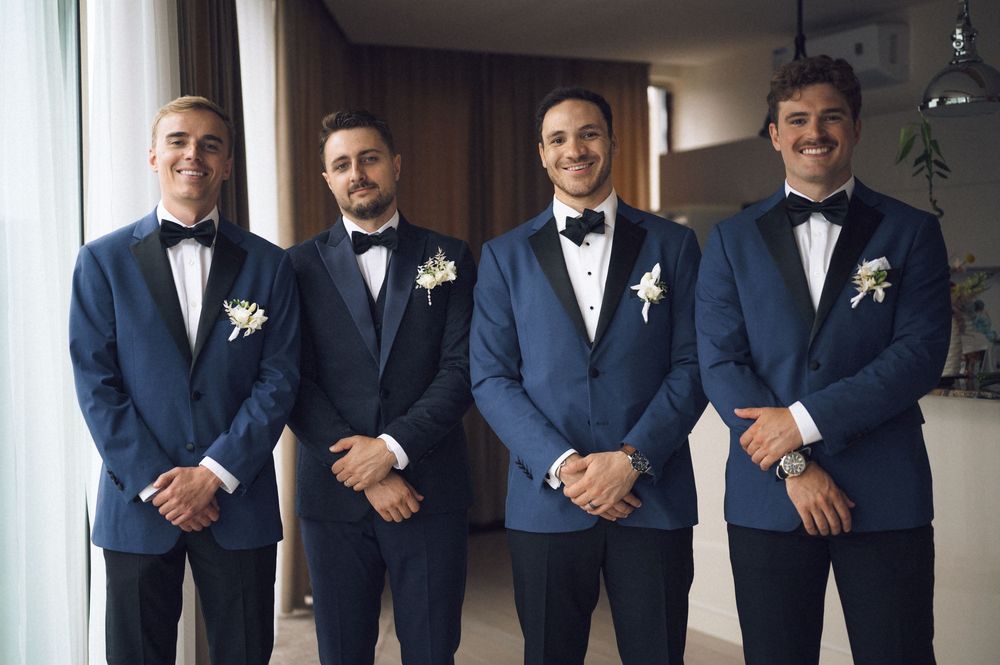 Four groomsmen in navy blue tuxedos with bow ties and white boutonnieres posing together at a wedding.